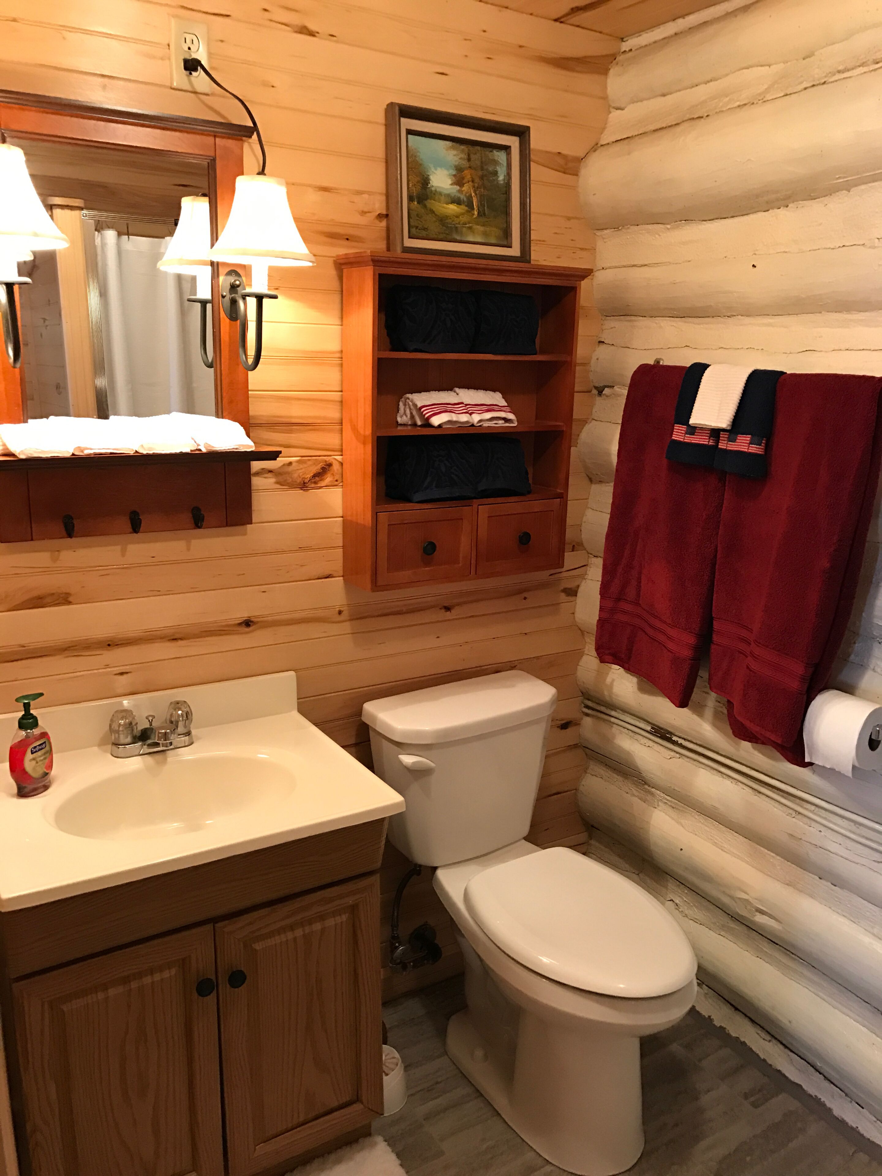 Un baño de cabaña de madera con un lavamanos, inodoro, estantes con toallas y un cuadro enmarcado en la pared.