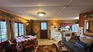 Ein gemütliches Blockhaushaus-Interieur mit Esstisch, Stühlen, einer Couch, einem Kamin, einer Küchenzeile und einem großen Fenster.