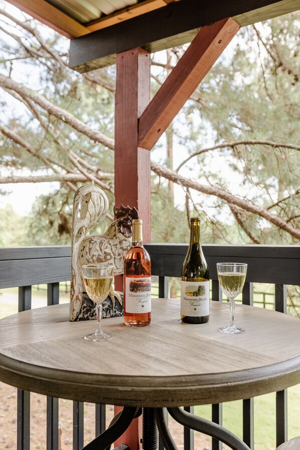 A round wooden table with two wine bottles and two glasses on it, set outdoors with a wooden railing and trees in the background.