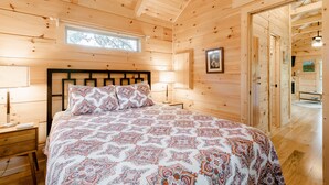 Une chambre à coucher dans une cabane en bois, comprenant un lit, des tables de chevet, une fenêtre et une porte donnant sur une autre pièce.