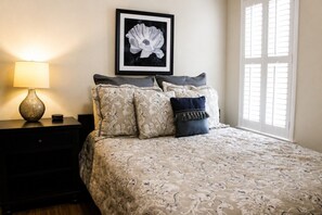 A bedroom with a bed, a nightstand, a lamp, a framed picture on the wall, and a window with blinds.