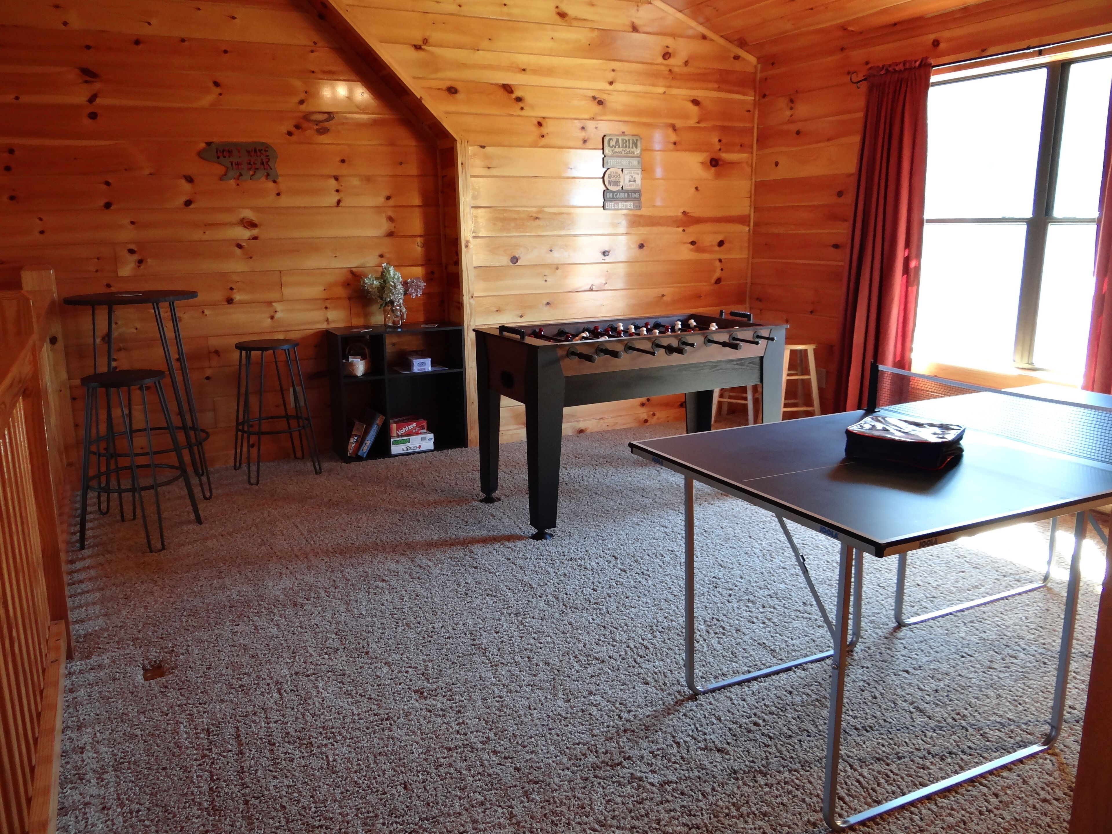 A cozy room with wooden walls, a foosball table, a ping pong table, and two stools.