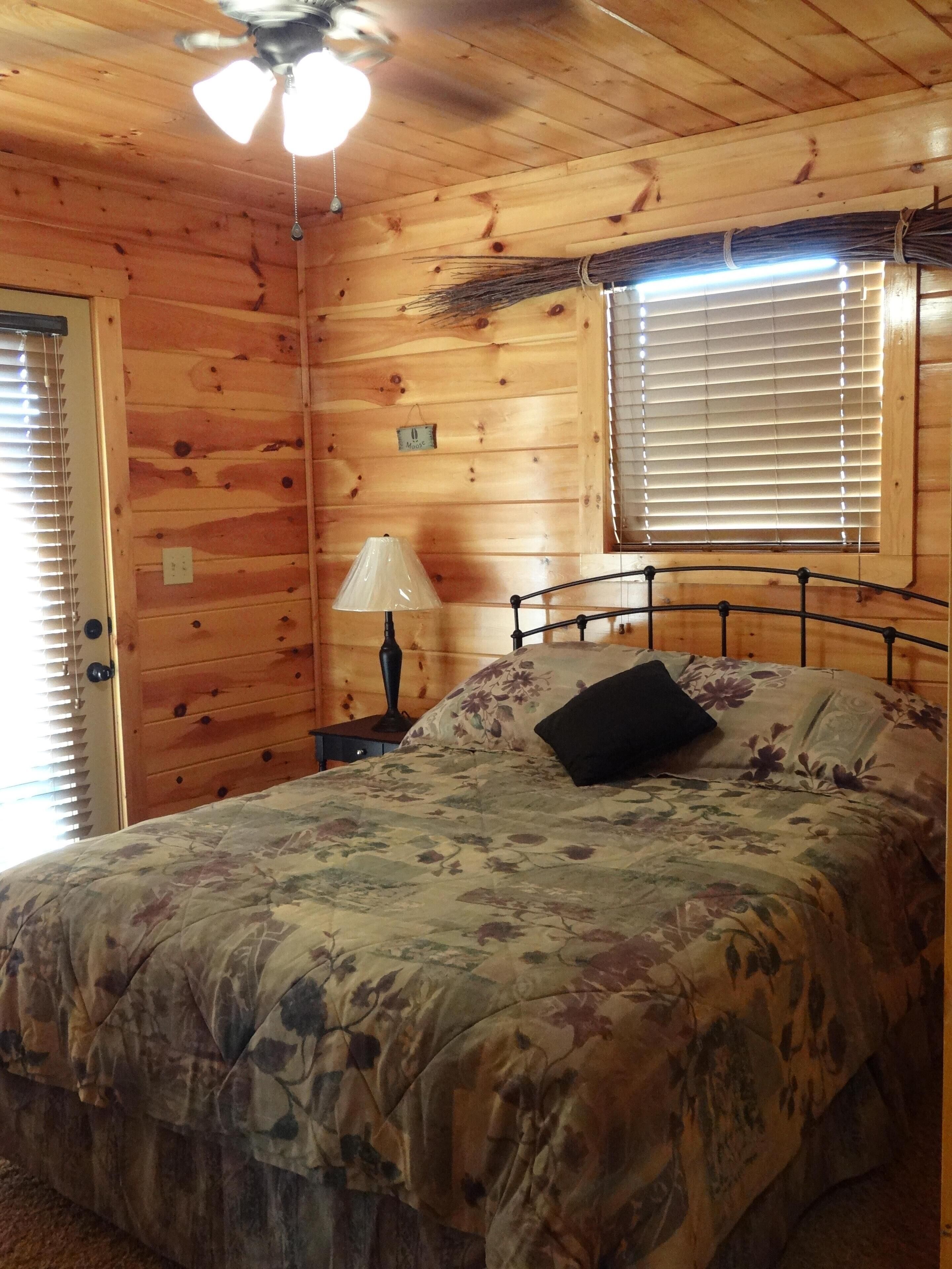 A wooden cabin bedroom with a bed, a nightstand, a lamp, a ceiling fan, and a window with blinds.