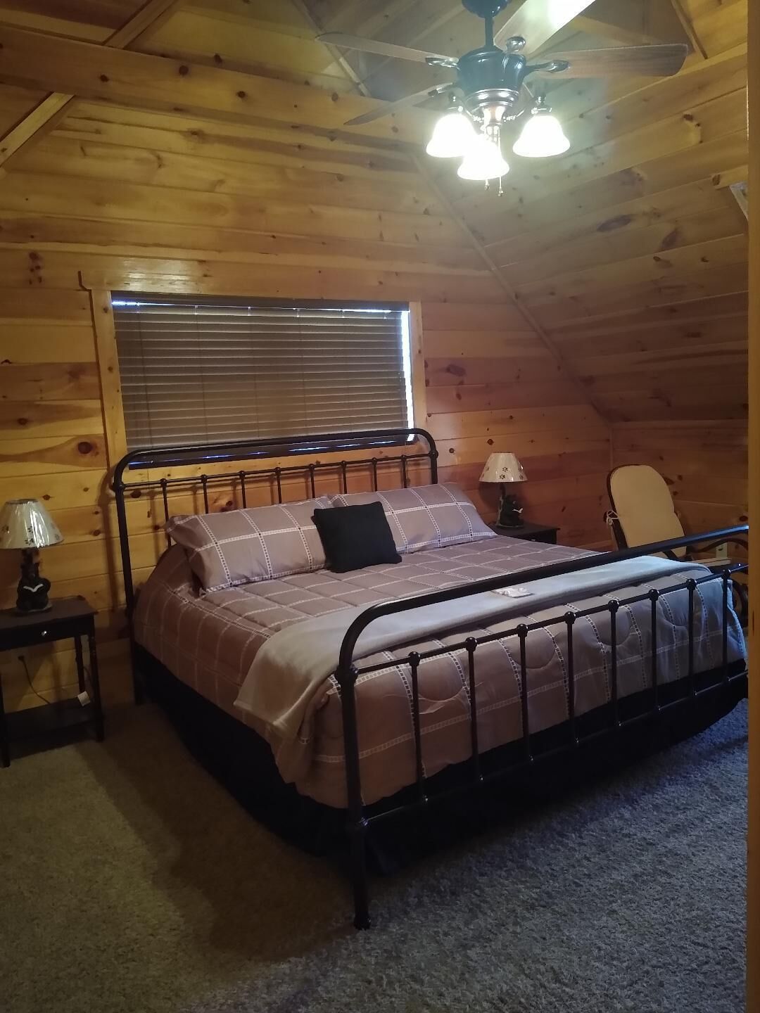 A wooden cabin bedroom with a metal bed frame, a ceiling fan, a window with blinds, a bedside table with a lamp, and a chair.