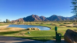 Un campo de golf con un estanque, montañas al fondo y un cielo azul despejado.