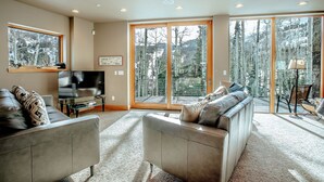A modern living room with a sofa, a television, and a large window overlooking a forested area.