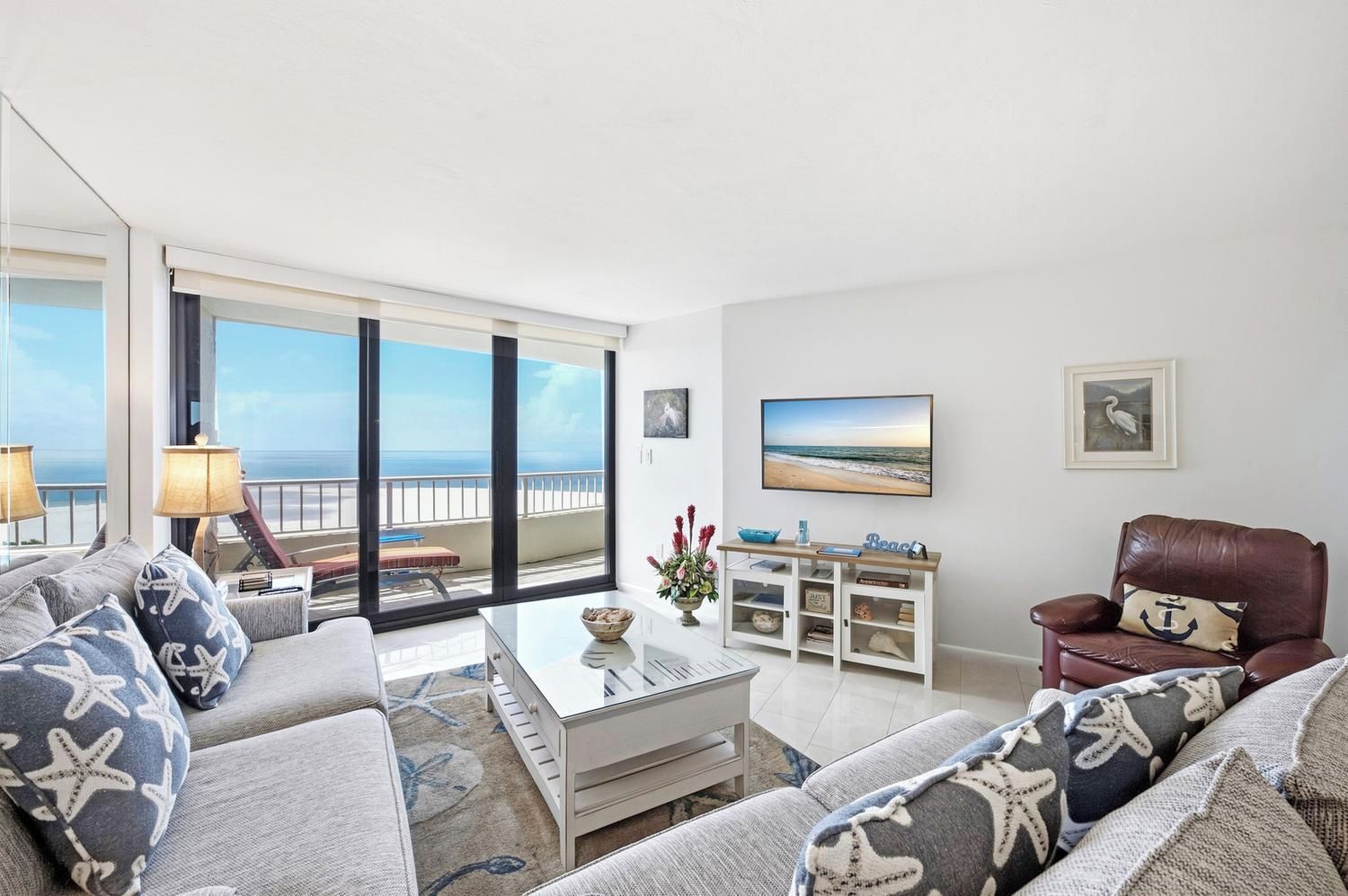 A spacious living room with a balcony overlooking the ocean, a comfortable sofa set, a coffee table, and a leather armchair.