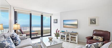 A spacious living room with a balcony overlooking the ocean, a comfortable sofa set, a coffee table, and a leather armchair.
