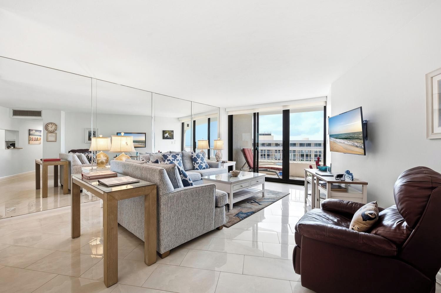 A spacious living room with a sofa set, a coffee table, and a leather armchair. There is a television mounted on the wall and a large window providing a view of the cityscape.