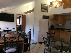 A compact kitchen with a refrigerator, wooden cabinets, a dining table with chairs, and a TV mounted on the wall.