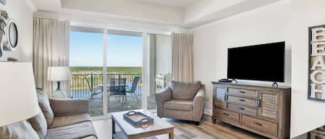 A spacious living room with a large flat-screen TV, a ceiling fan, and a view of the outdoors through a sliding glass door.