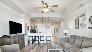 A modern living room with a flat-screen TV, a ceiling fan, and a kitchen area with bar stools. - Waterfront 1BR + Bunk Alcove at The Wharf • Sleeps 6 • Amazing Views • Oasis Pool (Orange Beach)