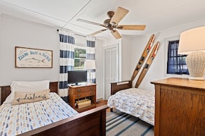 Quarto com duas camas, um ventilador de teto, uma placa emoldurada e uma janela com cortinas.