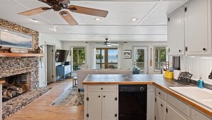 A spacious kitchen with a stone fireplace, wooden ceiling fan, and large windows offering a view of the outdoors.