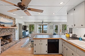 A spacious kitchen with a stone fireplace, wooden ceiling fan, and large windows offering a view of the outdoors.