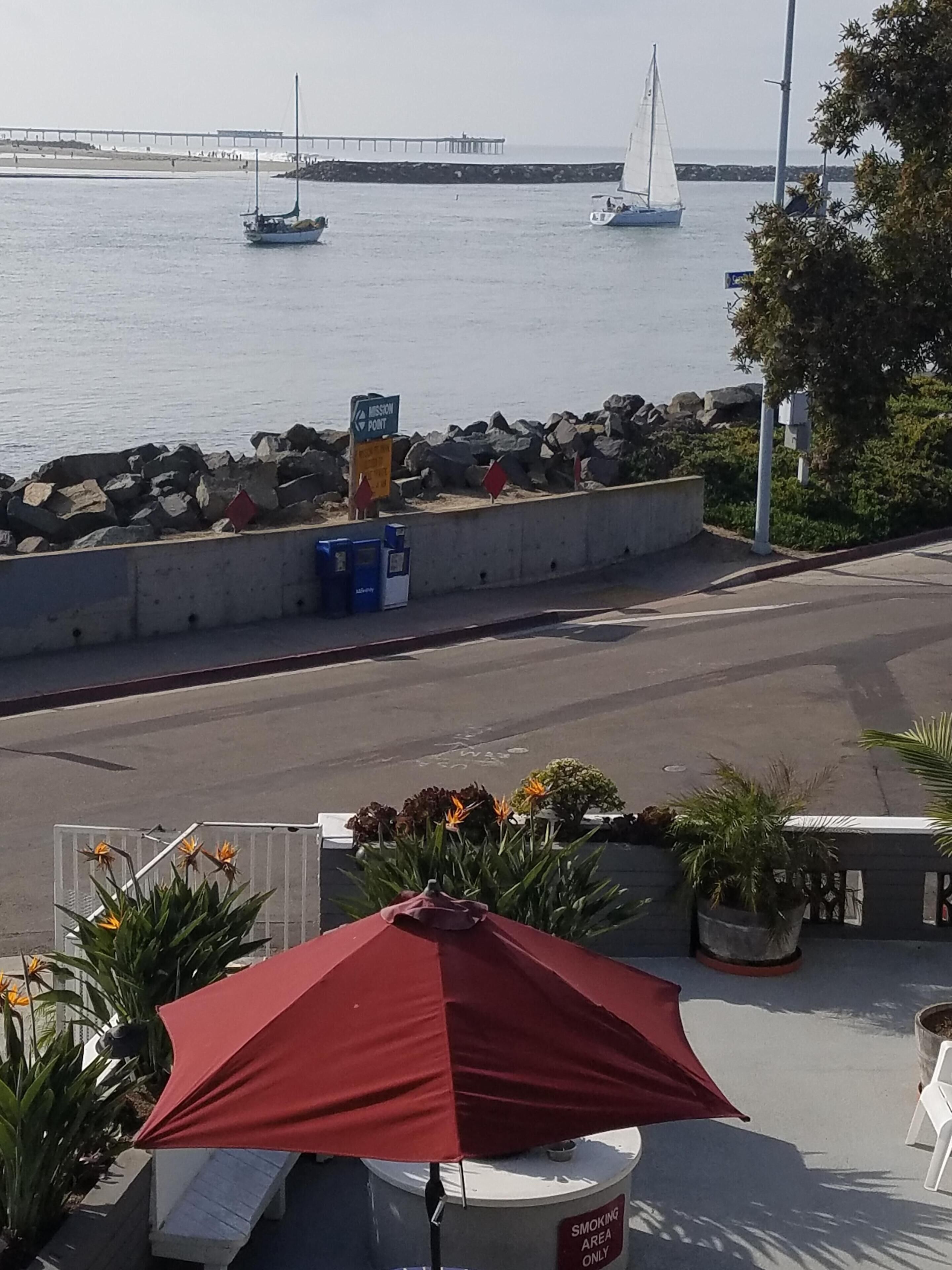 A seaside view with a rocky breakwater, a sailboat, and a bridge in the distance.