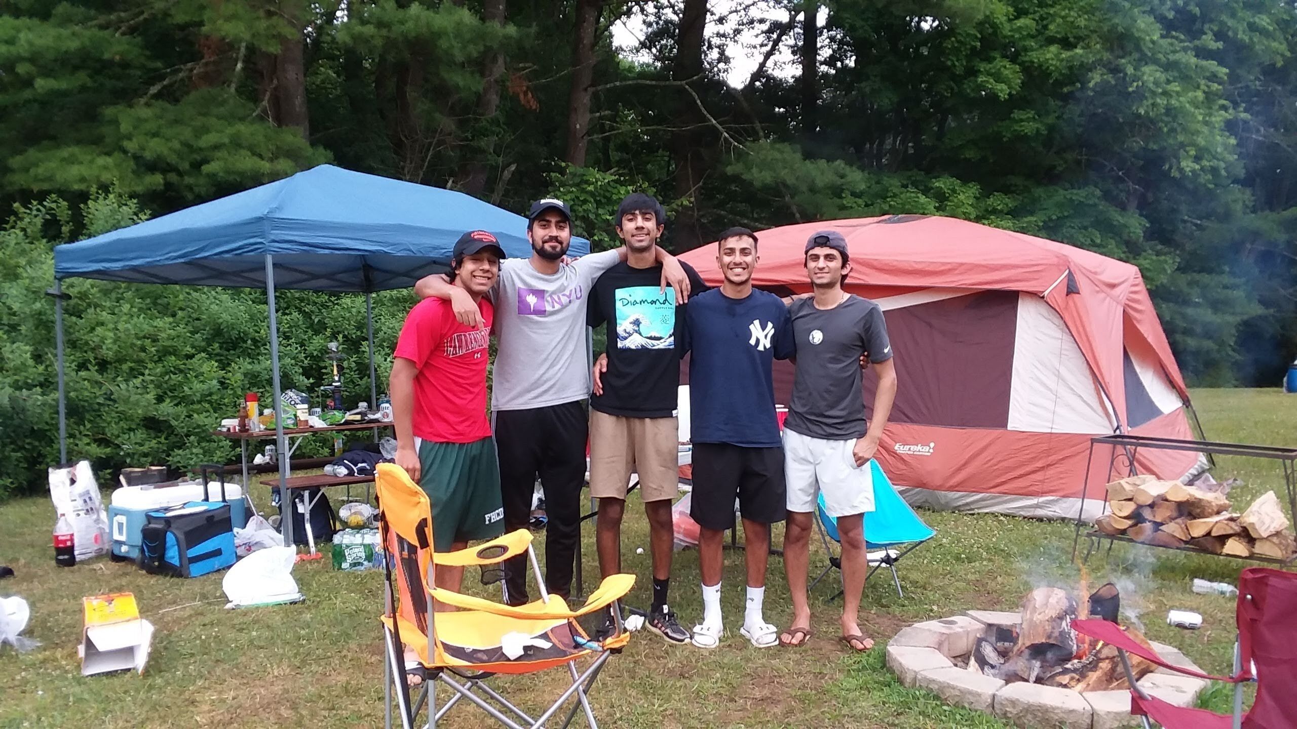 A group of people standing in front of a tent with a blue canopy and a red tent in a camping area.