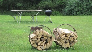 Une table de pique-nique, un barbecue et deux paniers en métal remplis de bois de chauffage, le tout installé dans un espace herbeux.