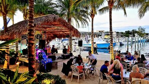 An outdoor dining area with tables and chairs, a stage with musical equipment, and a view of boats on the water. - POSHPADZ -Villa Koalua Walk to Jupiter Beach-Prime Lake Views-Resort Setting (Jupiter)