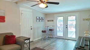 A living room with a ceiling fan, a sofa with a red pillow, a coffee table, and a bar area with a TV and remote controls. - Howdy Headquarters~ A Backyard Gathering Like no Other! (College Station)