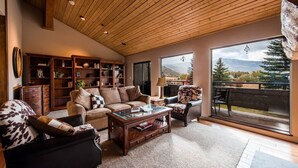 A spacious living room with a wooden ceiling, a large window with a mountain view, a leather sofa, a coffee table, and a bookshelf. - Wonderful Park City Townhome | Hot Tub | Mountain Views | Private Garage (Park City)