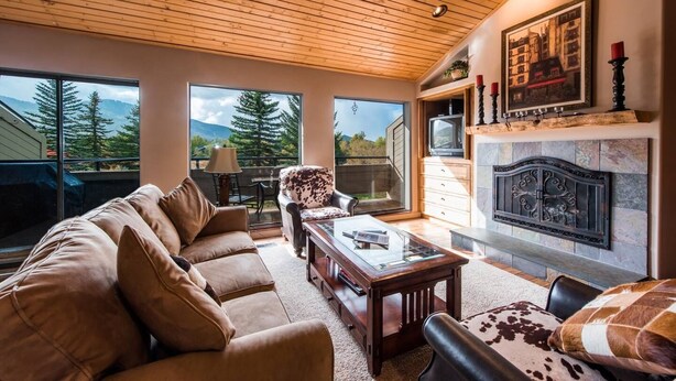 A spacious living room with a large sofa, a coffee table, and a fireplace. There are large windows offering a view of trees and mountains. - Wonderful Park City Townhome | Hot Tub | Mountain Views | Private Garage (Park City)