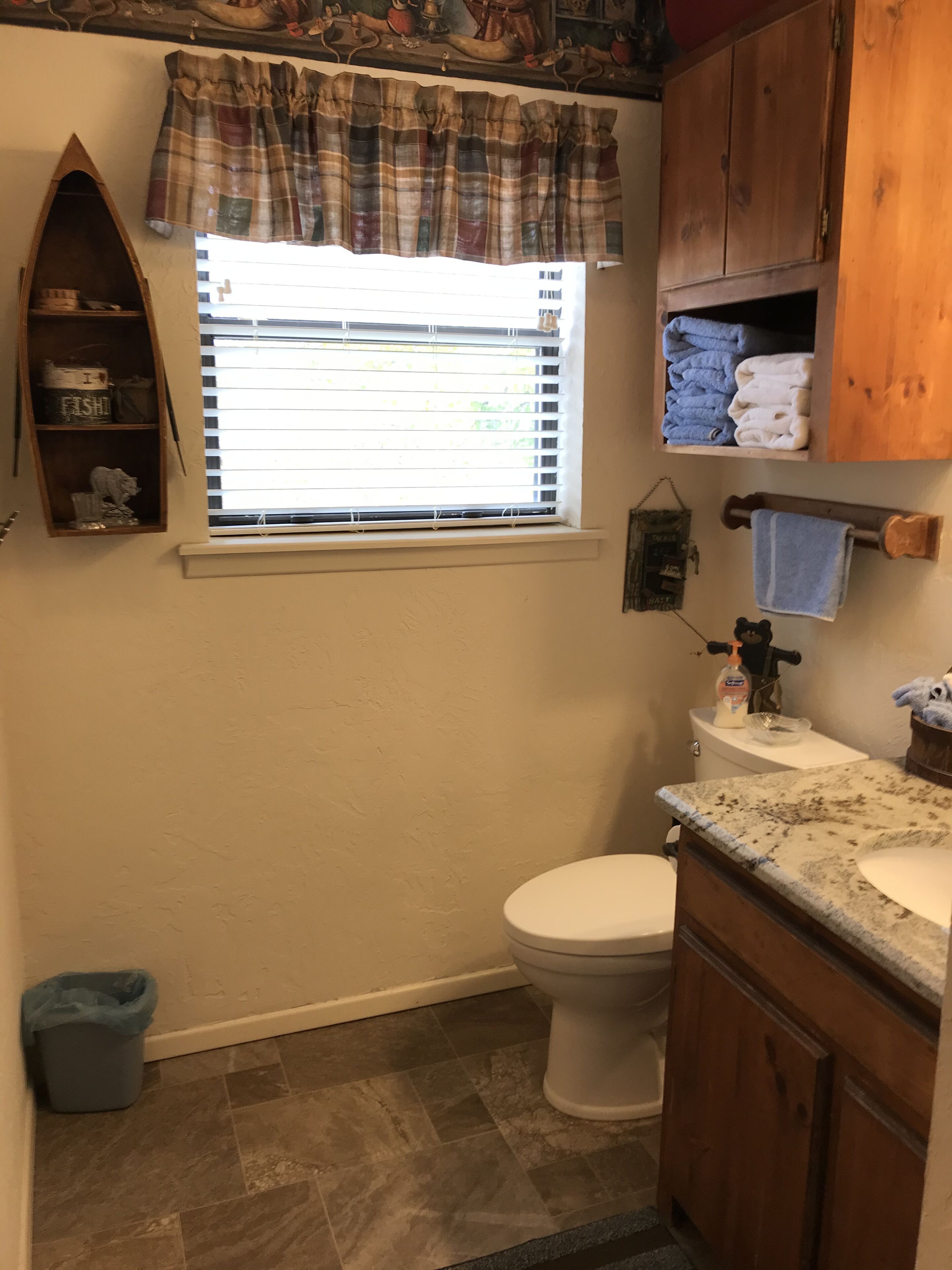 A bathroom with a toilet, a sink, a window with blinds, a shelf with towels, and a trash bin.