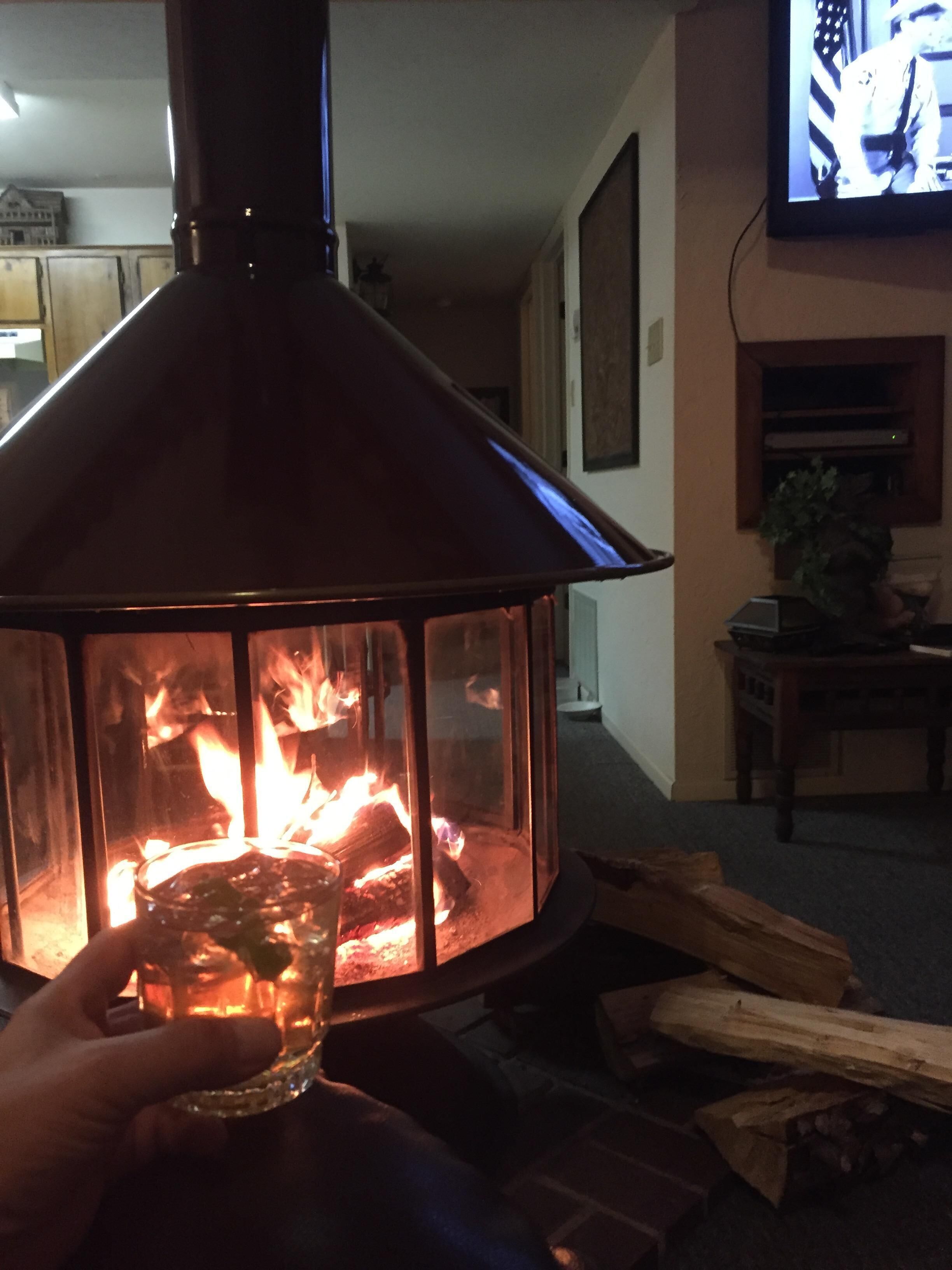 A fireplace with visible logs and flames, a television mounted on the wall, and a hand holding a glass.