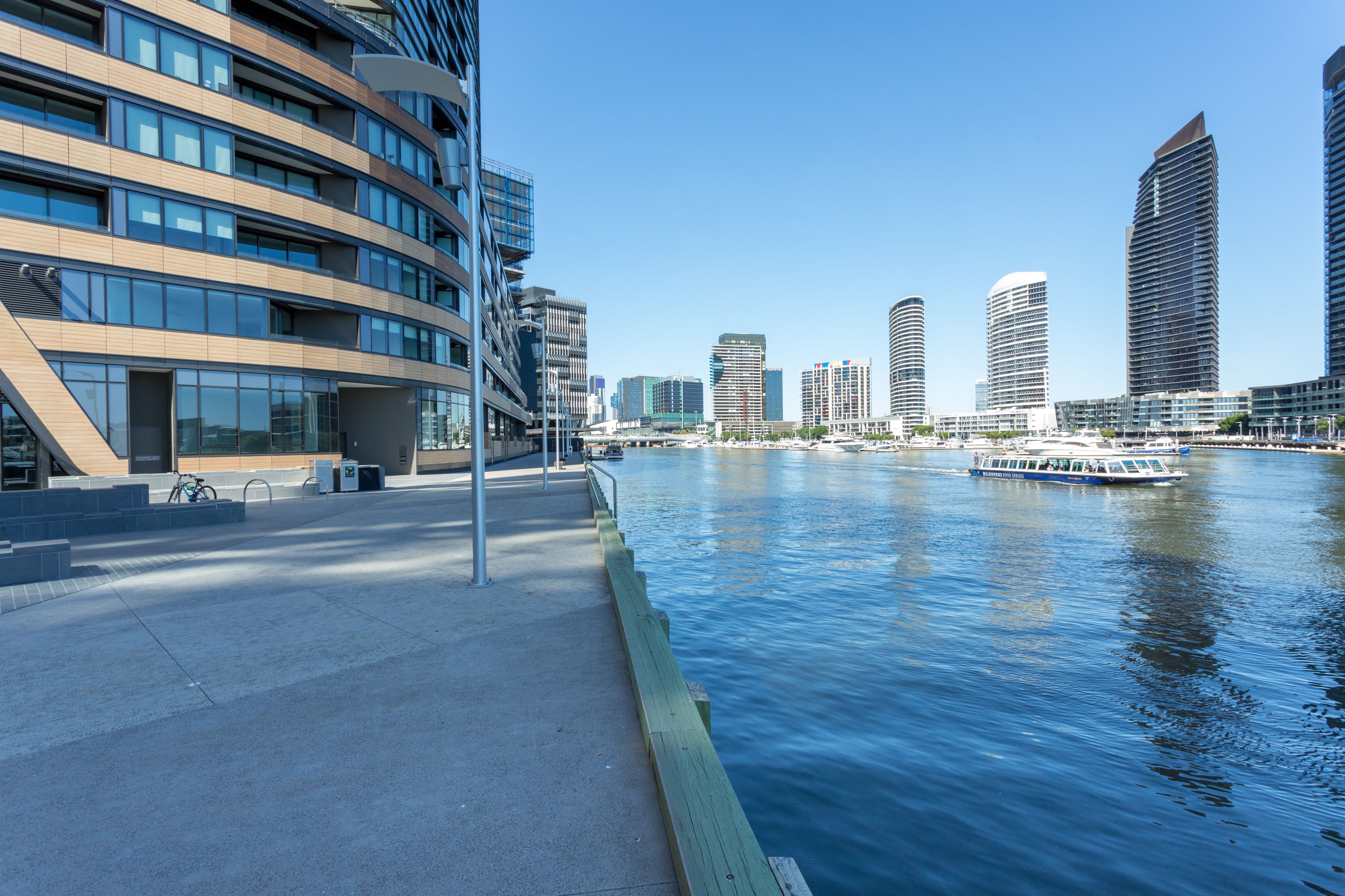A waterfront promenade with modern buildings, a calm river, and a boat docked on the right.