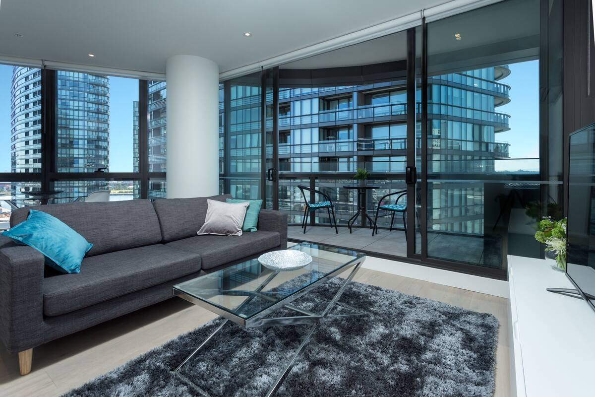 A modern living room with a grey sofa, glass coffee table, and a view of a high-rise building through large windows.
