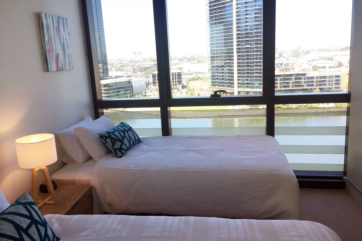 A hotel room with two beds, a nightstand with a lamp, and a large window offering a view of a cityscape and water.