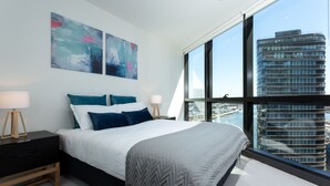 A modern bedroom with a large bed, two bedside tables, and a view of a cityscape through large windows. - Wyndel Apartments Riverside on Collins (Docklands)
