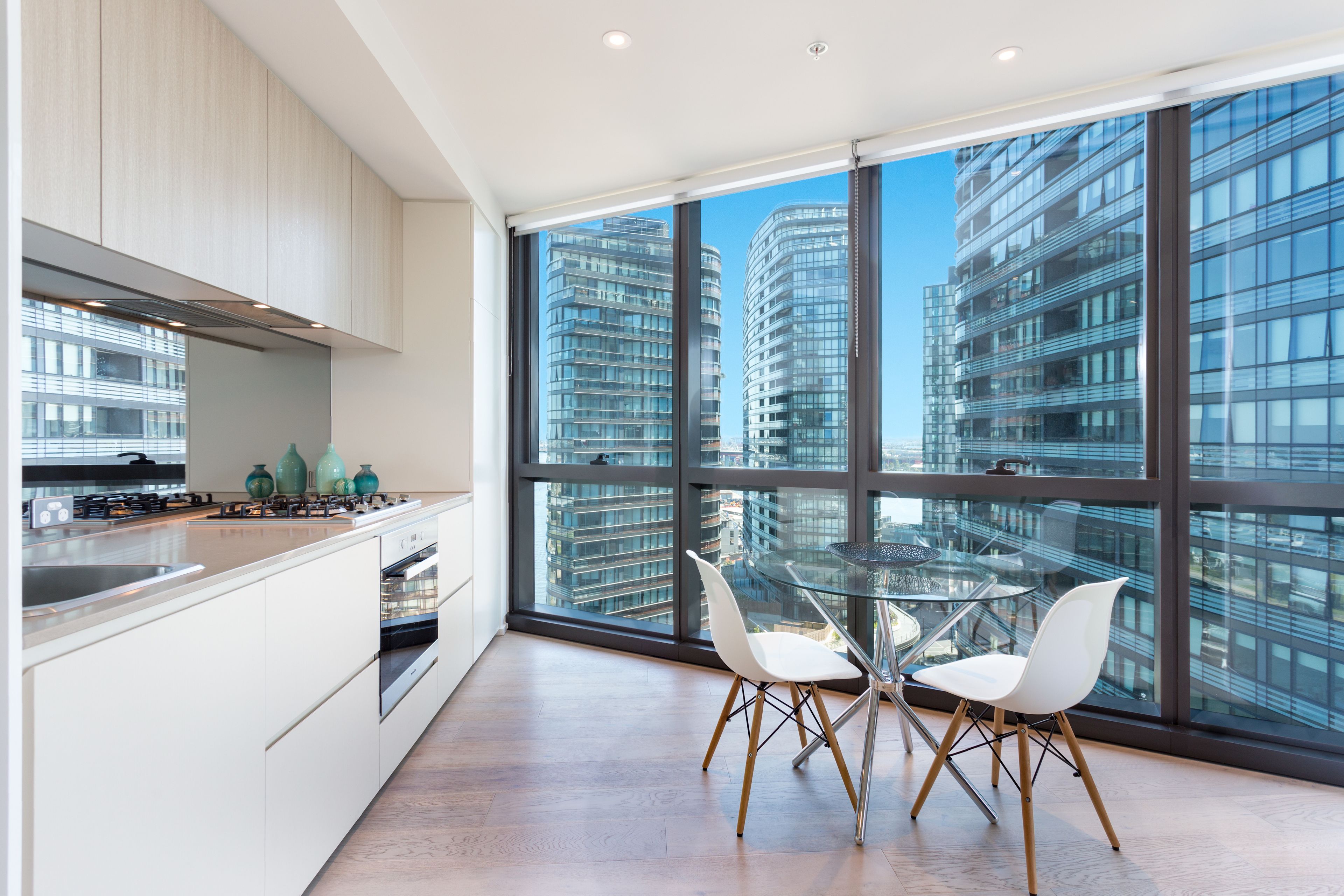 A modern kitchen with white cabinets, a glass dining table, and chairs, with a view of tall buildings through large windows.