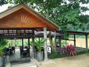 A covered outdoor seating area with tables and chairs, surrounded by potted plants and trees. - Balai Serama Guesthouse (Kuala Tembeling)