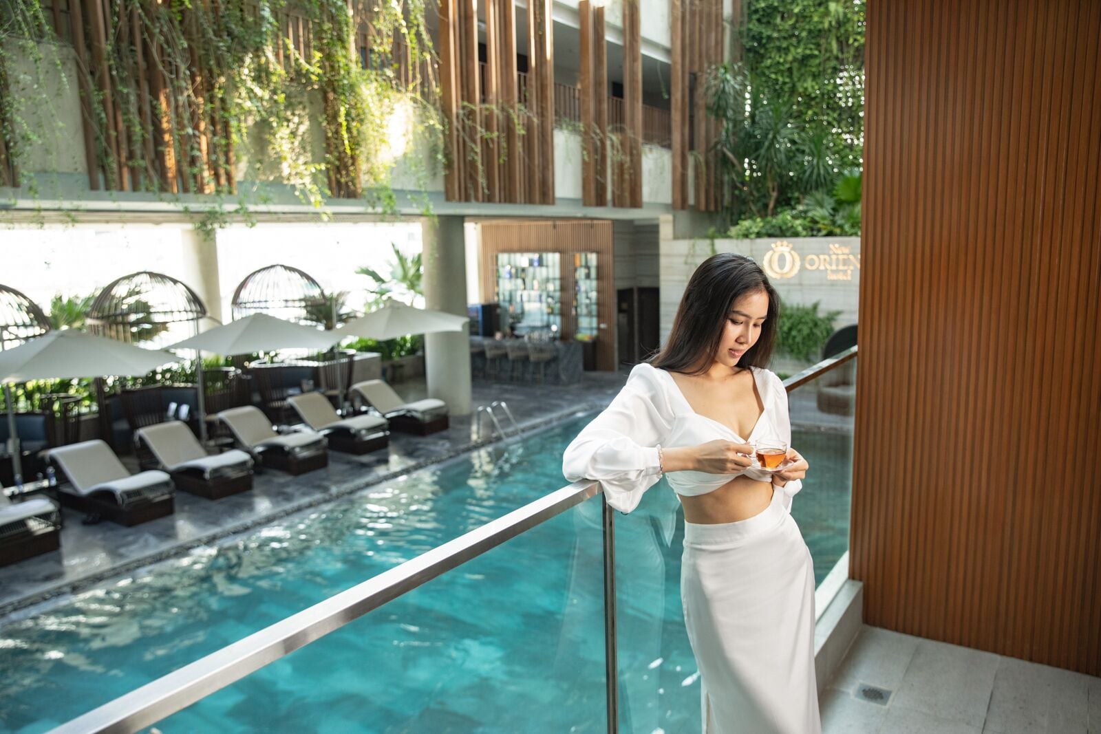 A woman in a white outfit by a pool with lounge chairs and umbrellas, a glass railing, and greenery in the background.