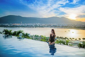 A woman sitting on an infinity pool with a cityscape and mountains in the background. - New Orient Hotel Danang (Da Nang)
