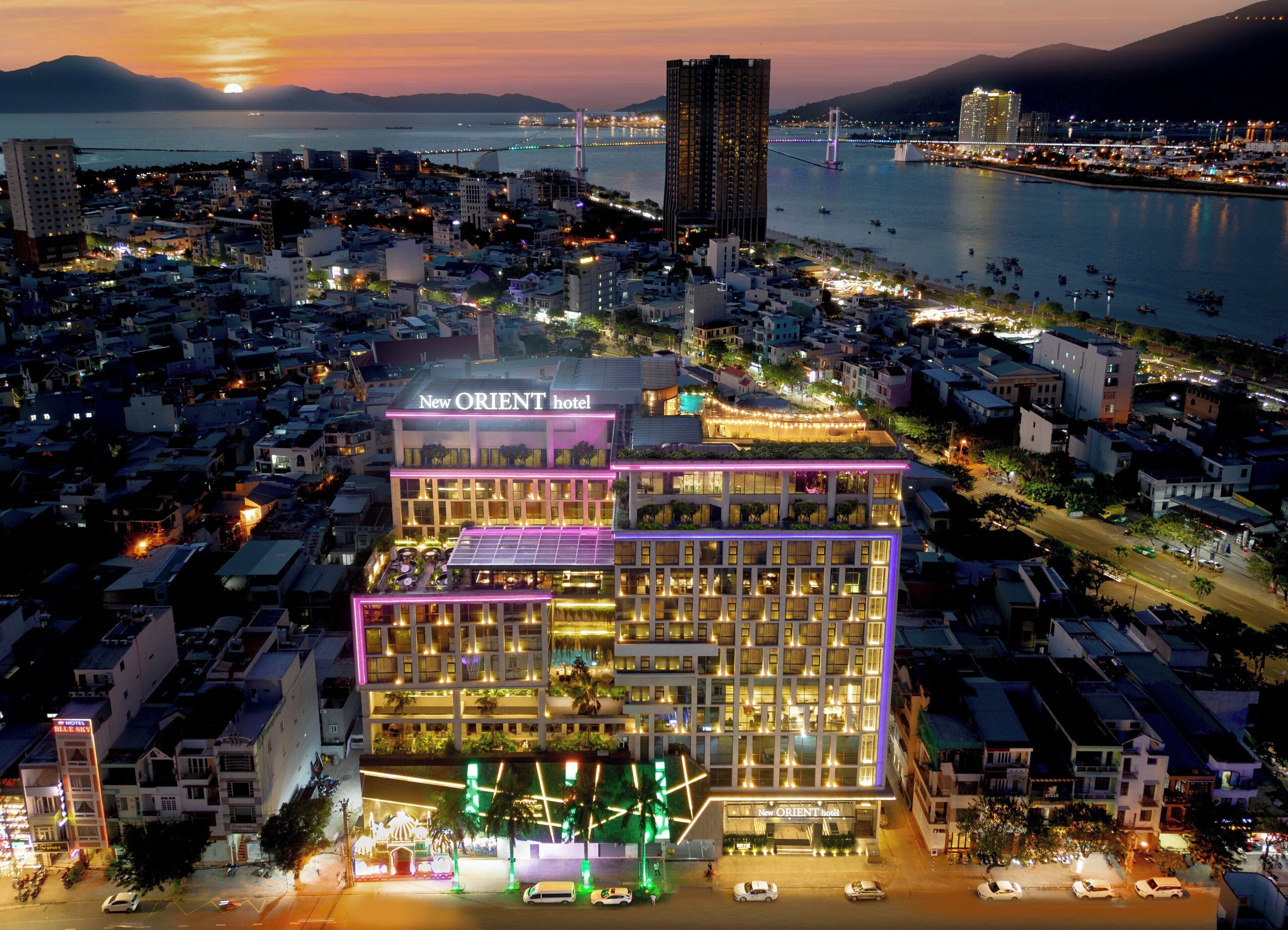 A modern hotel with multiple floors, a rooftop garden, and a view of the cityscape and water at dusk.