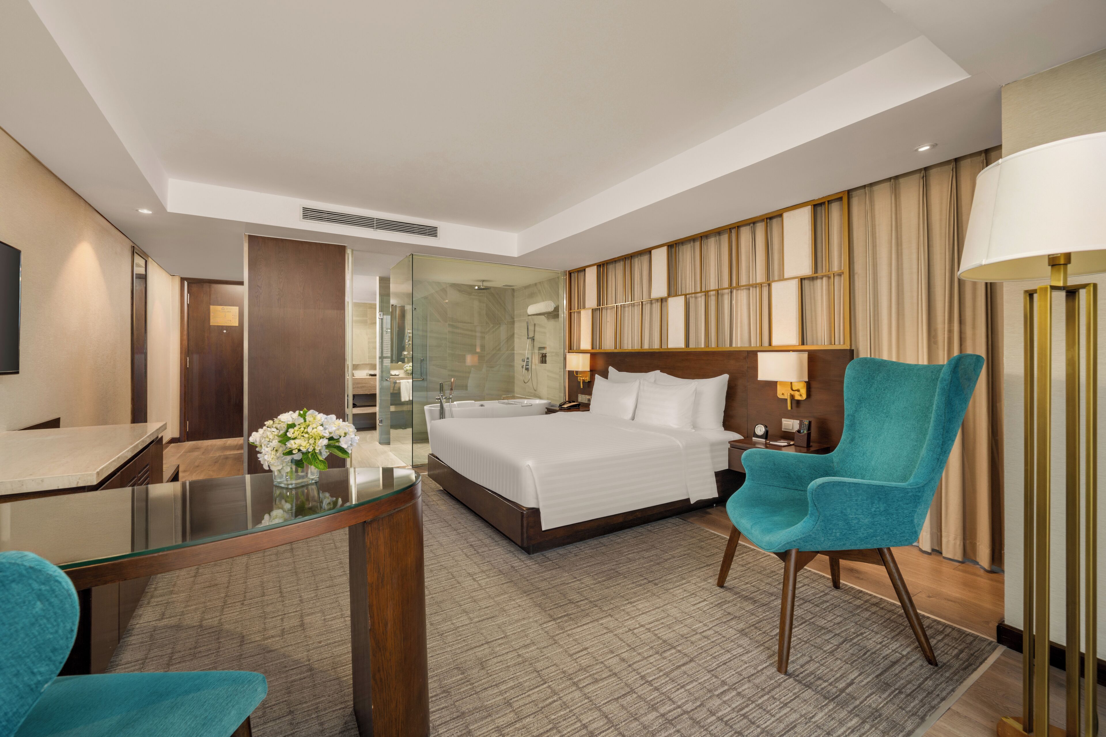 Ein Hotelzimmer mit einem großen Bett, einem Schreibtisch mit Glasplatte, einem blaugrünen Sessel und einer Lampe.