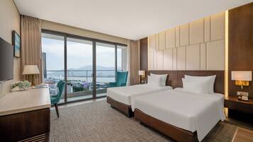 A hotel room with two beds, a desk, a chair, a TV, and a large window with a view of a cityscape and water.