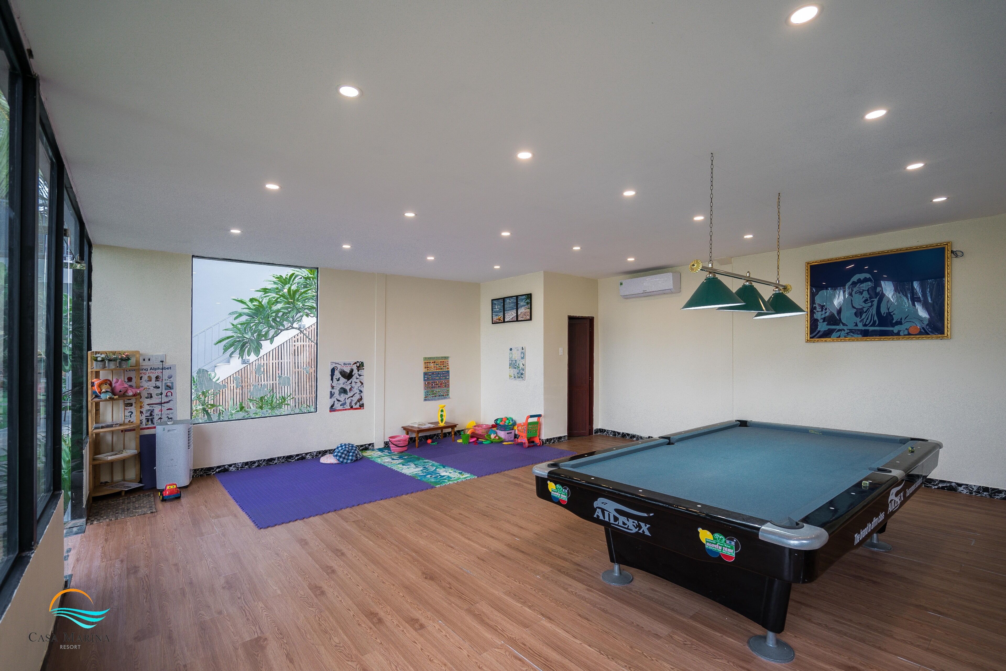 Una mesa de billar en una habitación con suelo de madera, un ventanal y vista a palmeras.