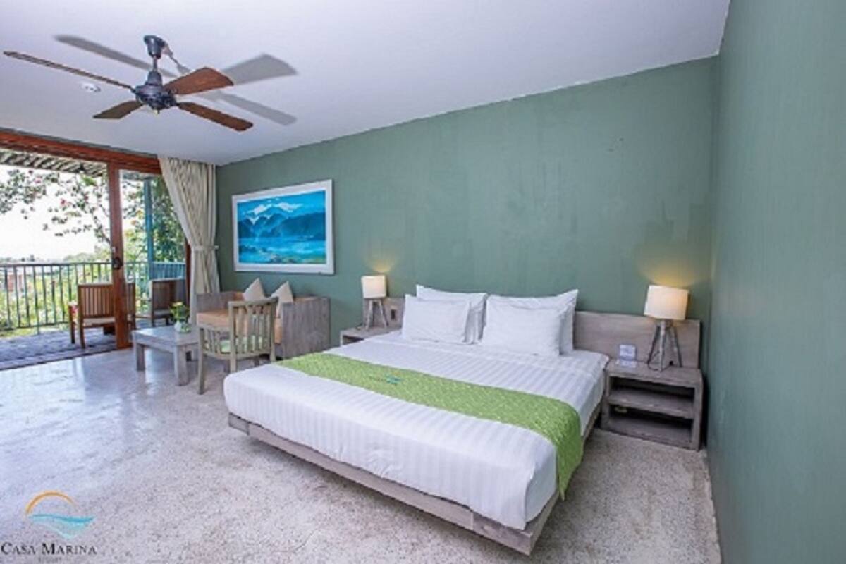 Ein Schlafzimmer mit einem Bett, zwei Nachttischen mit Lampen, einem Deckenventilator, einem Bild an der Wand und einem Balkon mit Stuhl und Tisch.