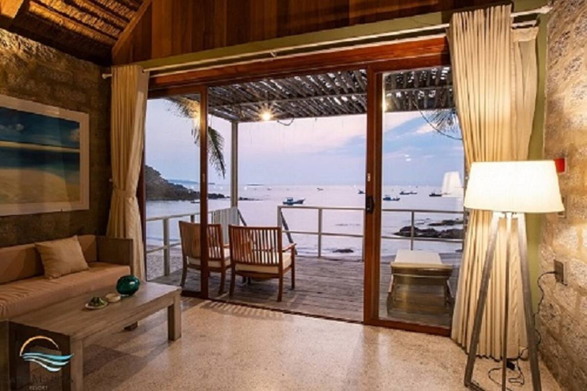 Ein Zimmer mit Holzbalkendecke, einer Couch, einem Couchtisch, einem Sessel, einer Lampe und Meerblick durch eine Glastür.