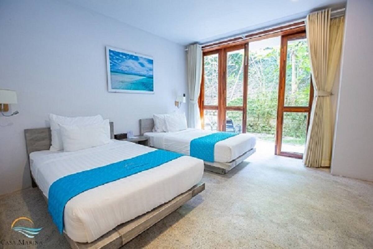 Ein Hotelzimmer mit zwei Betten, einem großen Fenster mit Vorhängen, einem Gemälde an der Wand und einem Logo auf dem Boden.