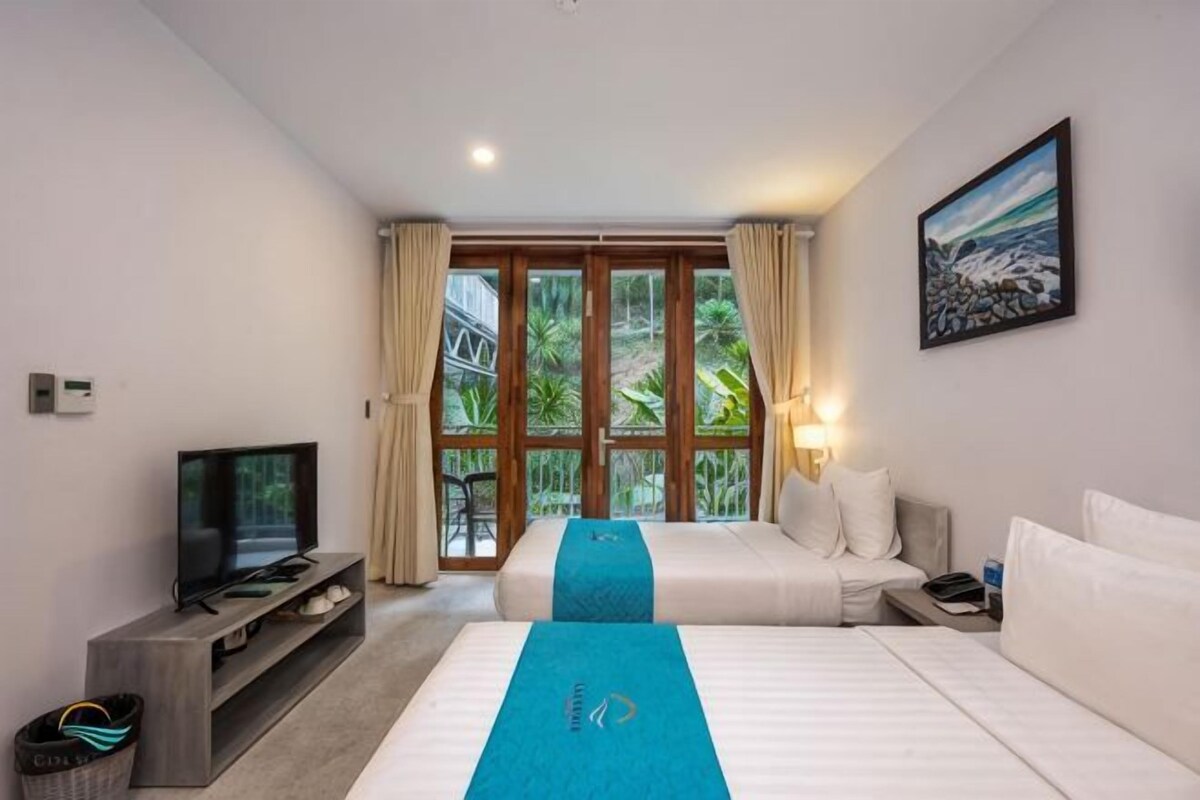 A hotel room with two beds, a TV, a desk, and a view of greenery through large windows.