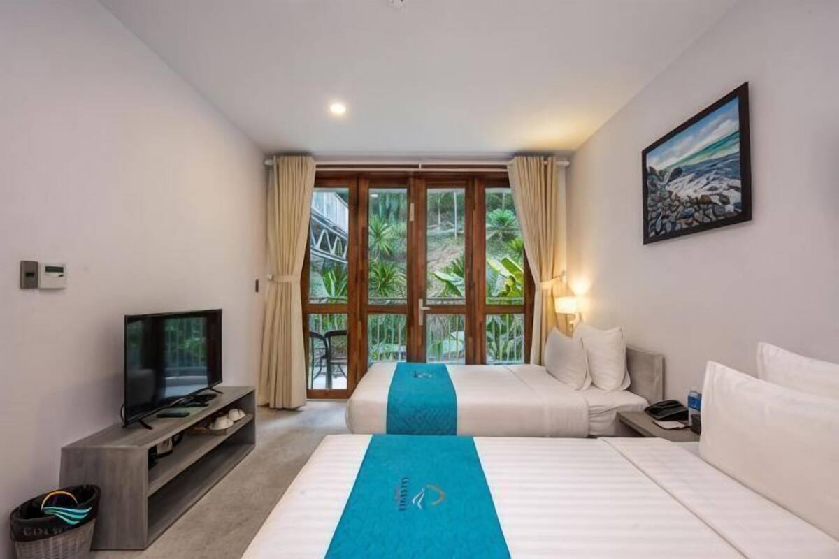 Ein Hotelzimmer mit zwei Betten, einem Fernseher, einem Schreibtisch und Blick auf Grün hinter großen Fenstern.