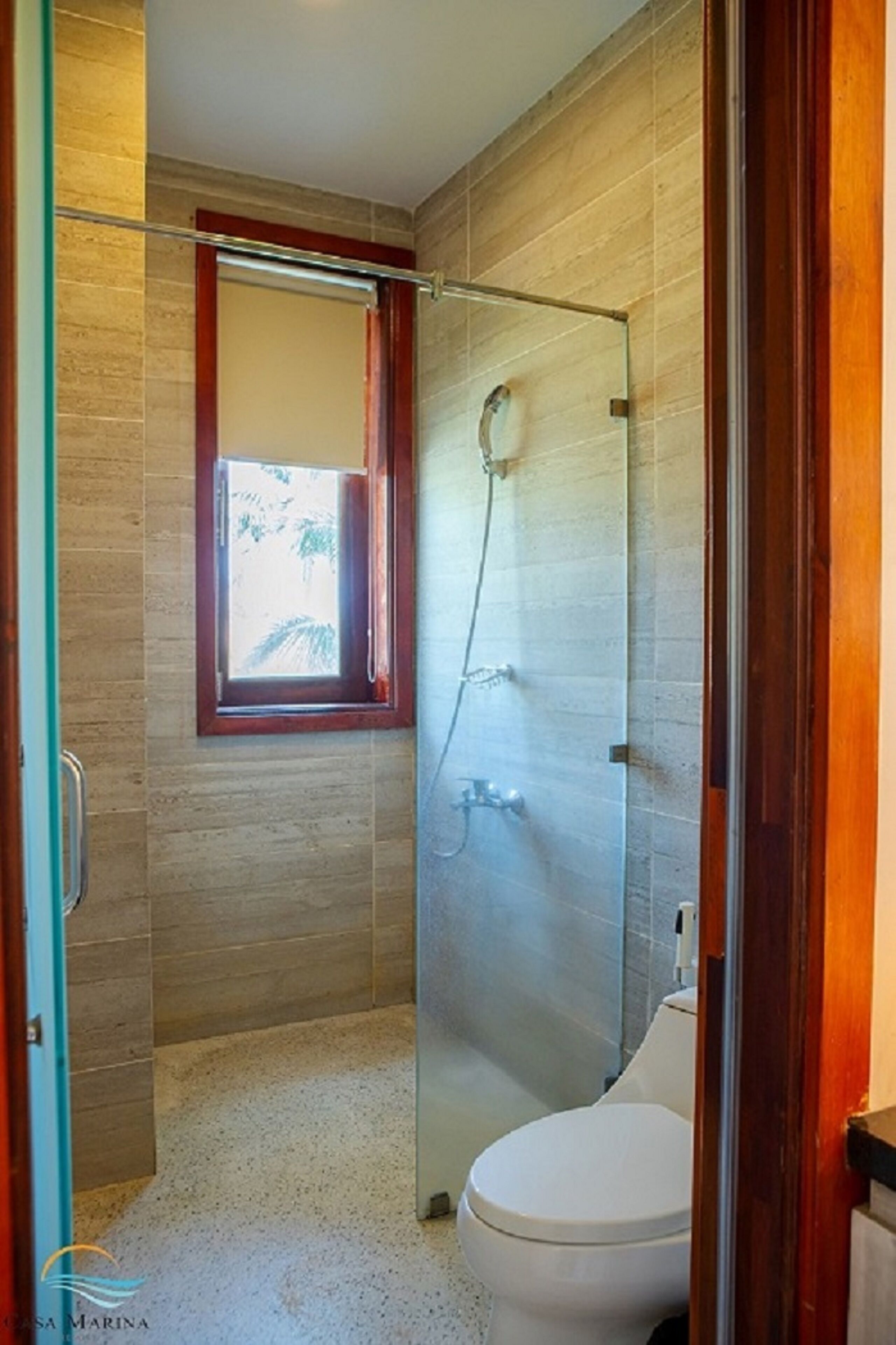 Un baño con ducha envuelta en vidrio, inodoro y ventana con vista a palmeras.