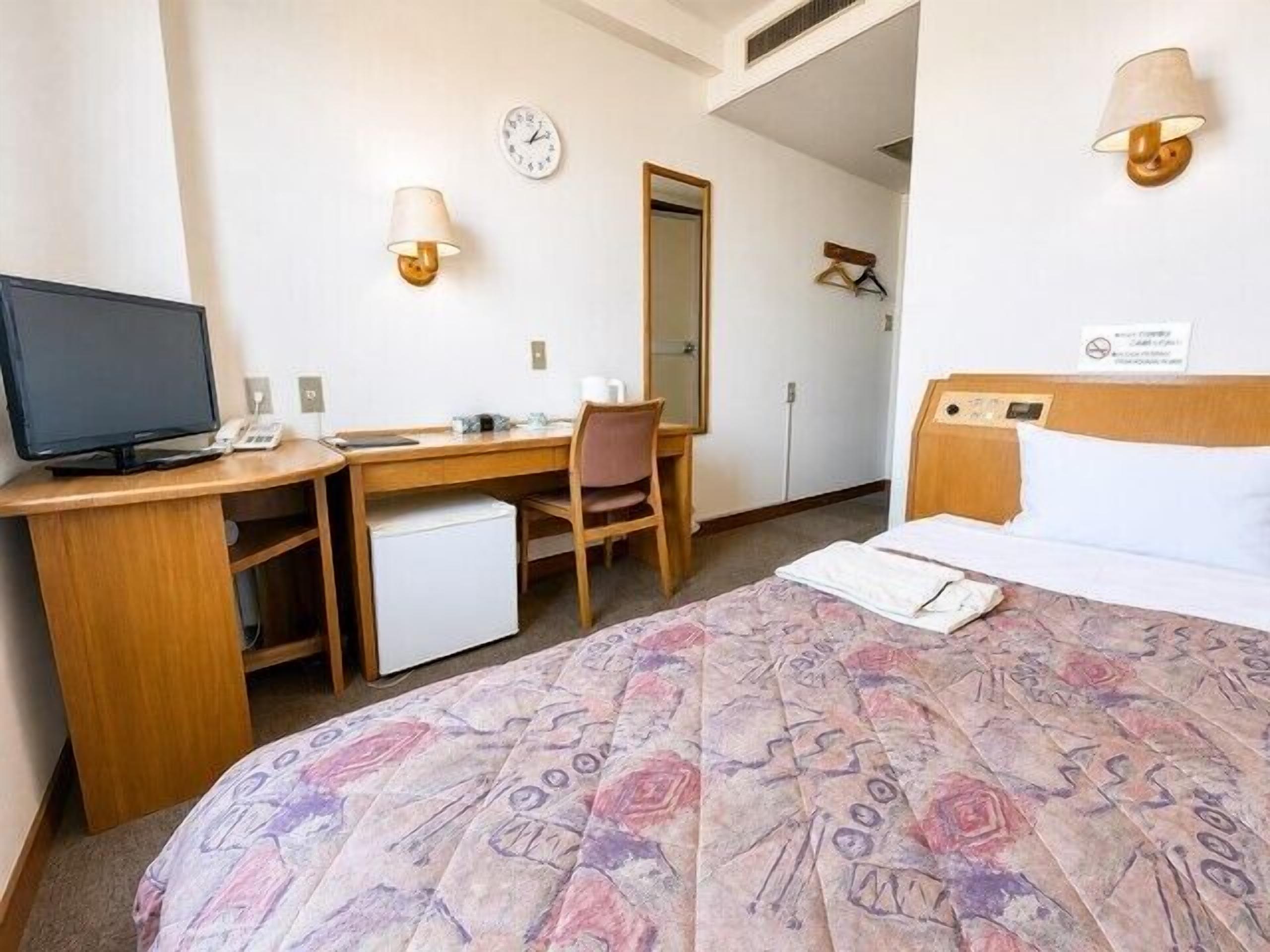 Een hotelkamer met een bed, bureau, stoel, televisie en een klok aan de muur.