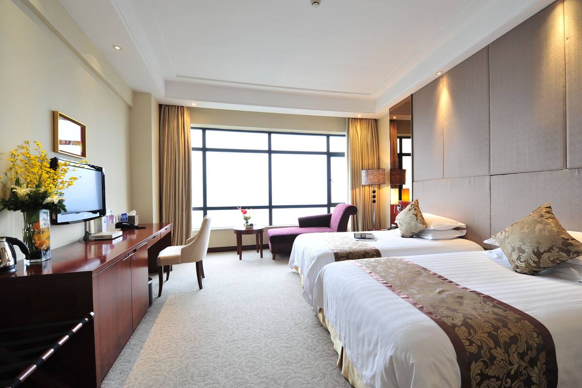A hotel room with two beds, a desk, a chair, a TV, and a large window.