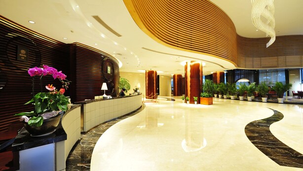A spacious lobby with a curved marble floor, wooden paneling, and a reception desk with potted plants. - Longting New Century Hotel Qiandao Lake (Hangzhou)