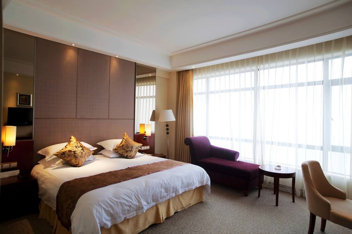 A hotel room with a large bed, a chair, a small table, and a large window with curtains.
